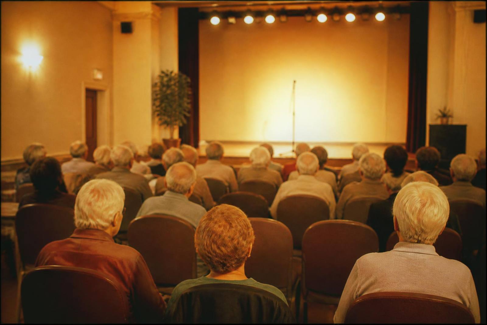 Residents seated from behind in an assisted living community room facing a small warmly lit stage