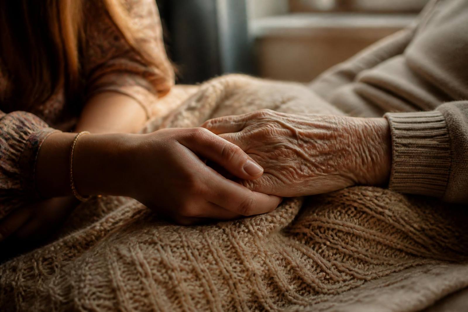 A daughter's hand resting on her mother's hand on a knitted blanket in soft window light, Orlando
